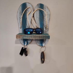 May include: A wooden wall-mounted key holder shaped like flip-flops. The flip-flops are painted blue and white with rope straps. A pair of sunglasses rests on a small shelf, and keys hang from hooks below.