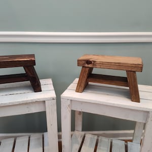 Op de afbeelding: Twee kleine houten krukjes met rechthoekige zittingen en schuine poten. Het ene krukje heeft een donkerbruine beits, terwijl het andere een lichtere, natuurlijke houtafwerking heeft. Beide krukjes hebben een eenvoudige, stevige constructie met zichtbare houtnerf.