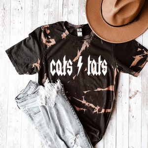 May include: A black tie-dye t-shirt with the text "cats & tats" in white block letters. The shirt is paired with light blue distressed jeans and a brown fedora.