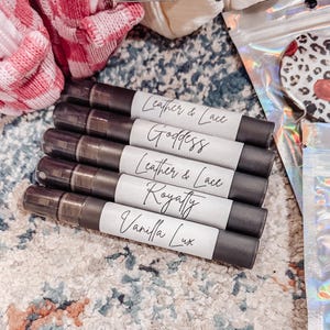 May include: A collection of five dark gray tubes with white labels. The labels feature handwritten text: "Leather & Lace", "Goddess", "Leather & Lace", "Royalty", and "Vanilla Lux". The tubes are arranged on a patterned surface, with other items in the background.