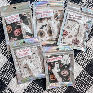 May include: Five small clear plastic bags with white fabric squares inside. Each bag contains a small clear bottle with a white liquid and a fabric square with a different Halloween-themed design. The designs include a pumpkin, ghosts, and sunflowers. The text on the bags reads "Farmhouse Pumpkin", "Apple Pumpkin Butter", "Vanilla Pumpkin Marshmallow", "Pumpkin Bread", and "Pumpkin Spice".