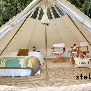 May include: A white canvas tent with a large window in the roof. The tent is set up inside a forest and has a bed, two chairs, a table, and a lantern inside. The tent has a rug on the floor and a tote bag with the word "Fernweh" printed on it.