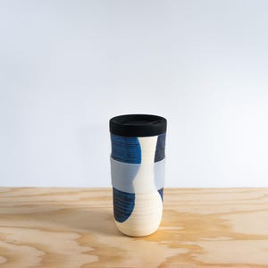 Könnte beinhalten: Ein Keramik-Reisebecher mit schwarzem Deckel und einem hellgrauen Silikonband. Der Becher hat ein blau-weißes abstraktes Design. Der Becher ist zylindrisch mit abgerundetem Boden und steht auf einer Holzoberfläche.