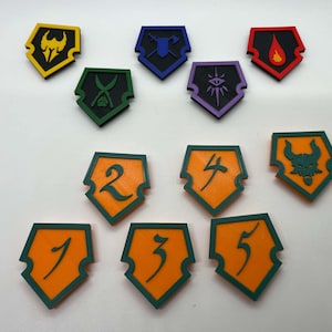 May include: A set of 10 colorful, shield-shaped tokens with various symbols and numbers. The tokens are made of plastic and have a green border. The symbols include a sword and axe, a flame, a dragon head, and an eye. The numbers are 1 through 5.