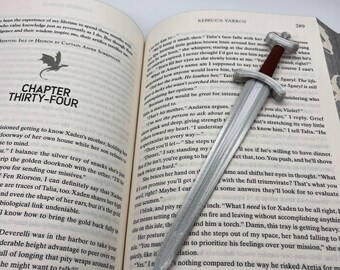 Puede incluir: Un libro blanco con un marcapáginas de espada plateada. El libro está abierto en una página con el título "Capítulo Treinta y Cuatro" y texto sobre una madre y su hijo. El marcapáginas de espada es un accesorio decorativo.