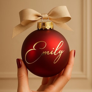 Könnte beinhalten: Eine dunkelrote Weihnachtskugel mit dem Namen "Emily" in goldener Schrift. Sie hat eine goldene Kappe und eine goldene Schleife. Die Kugel wird in einer Hand gehalten.