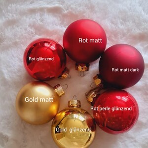 Pu&ograve; includere: Una collezione di decorazioni natalizie in varie tonalit&agrave; di rosso e oro. Le decorazioni includono finiture opache e lucide, con etichette di testo in tedesco. Le decorazioni sono disposte a cerchio.