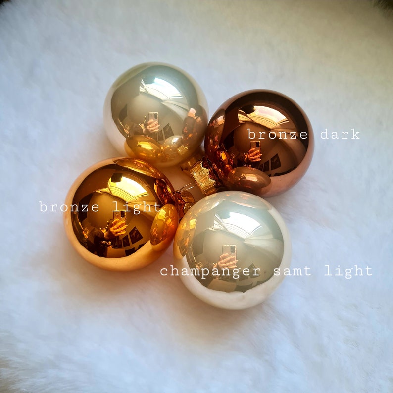 Pu&ograve; includere: Quattro sfere natalizie riflettenti, in tonalit&agrave; bronzo e champagne. Le sfere sono etichettate come "bronze light", "bronze dark" e "champanger samt light". Sono disposte su una superficie bianca e soffice.