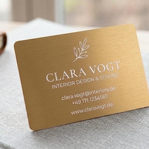 Könnte beinhalten: Eine goldene Visitenkarte mit dem Namen "CLARA VOGT" und Kontaktinformationen. Die Karte hat ein Blattdesign und liegt auf einer grauen Oberfläche mit einem Notizbuch, Stift und Vase im Hintergrund.