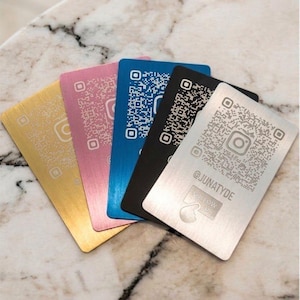 Può includere: Un set di cinque carte rettangolari in metallo color oro, rosa, blu, nero e argento. Ogni carta presenta un codice QR inciso, un logo Instagram, il testo @JUNATYDE e un'icona a forma di mano con il testo FOLLOW US sulla superficie.