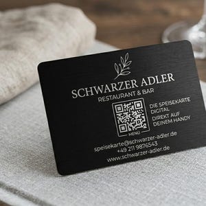 Könnte beinhalten: Eine schwarze Visitenkarte mit weißem Text "SCHWARZER ADLER RESTAURANT & BAR". Sie enthält einen QR-Code und Kontaktdaten. Die Karte liegt auf einer grauen Oberfläche neben einer Leinen-Serviette.