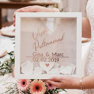 May include: A square white shadow box frame containing a light background with the text "Unser Polterabend", "Gina & Marc", and "02.02.2019" in copper lettering. The bottom of the frame is filled with small white paper hearts.