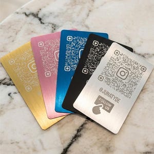 Könnte beinhalten: Eine Sammlung metallischer Visitenkarten in Gold, Rosa, Blau, Schwarz und Silber. Jede Karte zeigt einen QR-Code und ein Instagram-Logo. Die silberne Karte hat den Text "@JUNATYDE FOLLOW US".