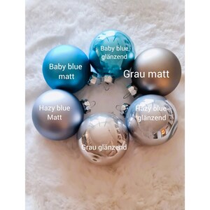 Pu&ograve; includere: Sei palline di Natale in varie tonalit&agrave; di blu e grigio, con finiture opache e lucide. Le etichette includono "Baby blue matt" e "Grau gl&auml;nzend". Le palline sono disposte su una superficie bianca strutturata.