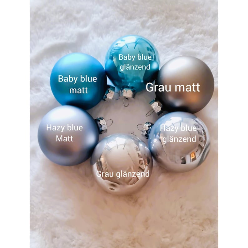 Pu&ograve; includere: Sei palline di Natale in varie tonalit&agrave; di blu e grigio, con finiture opache e lucide. Le etichette includono "Baby blue matt" e "Grau gl&auml;nzend". Le palline sono disposte su una superficie bianca strutturata.