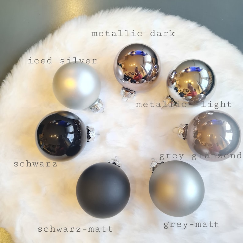 Pu&ograve; includere: Una collezione di palline di Natale in varie tonalit&agrave; di grigio e argento, con finiture opache e metalliche. Il testo sull'immagine indica i colori: "iced silver", "metallic dark", "metallic light", "grey gl&auml;nzend", "schwarz", "schwarz-matt" e "grey-matt".