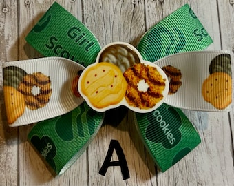 Könnte beinhalten: Grün-weißer Girl Scout Cookie-Bogen mit verschiedenen Keksen auf dem weißen Band. Die Schleife ist mit einem grünen Band gebunden, auf dem "Girl Scout Cookies" steht.