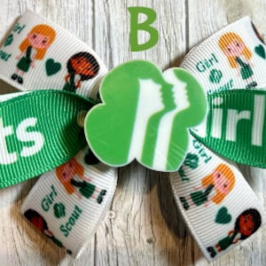 May include: Green and white Girl Scout bow with a green clover-shaped charm with the Girl Scout logo. The bow has the words "Girl Scout" and "Girl Scouts" printed on it.
