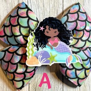 May include: A black and white mermaid hair bow with a mermaid with black hair and a pink top. The bow is made of a fabric with a mermaid scale pattern.
