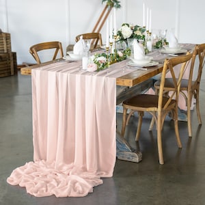 May include: A blush pink chiffon table runner with a subtle ombre effect, draped over a rustic wooden table with a floral centerpiece and candles.
