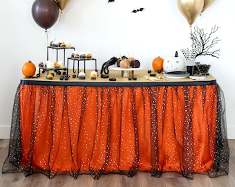 May include: Orange and black Halloween party table setting with a black tulle table skirt, black bats, and balloons. There are cupcakes, candy, and a cake on the table.