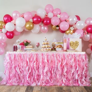 May include: A pink and white balloon arch with pink and gold accents decorates a table with a pink ruffled tablecloth. The table is set with a variety of desserts, including cupcakes, donuts, and strawberries.