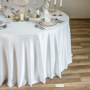 May include: A round, white satin tablecloth covers a table set for a formal event. The table is adorned with floral centerpieces, candles, and elegant place settings. The tablecloth drapes gracefully to the floor, creating a luxurious look.