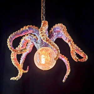 May include: A unique octopus-shaped chandelier with a warm, glowing light. The tentacles are a mix of purple, orange, and gold, and they surround a spherical light fixture. The fixture hangs from a chain against a dark background.