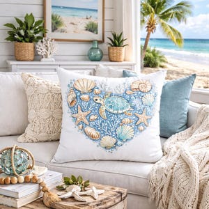 Pode incluir: Uma almofada decorativa quadrada branca com uma ilustração central em forma de coração composta por várias conchas, corais e uma tartaruga marinha azul-clara. O design utiliza tons de azul, bege e branco para um motivo temático marinho.