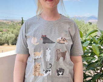 May include: A gray t-shirt featuring a collection of cartoon cat illustrations in various poses and colors. The cats include a black cat with a red tongue, a calico cat curled up, and other breeds. The shirt is being worn outdoors.