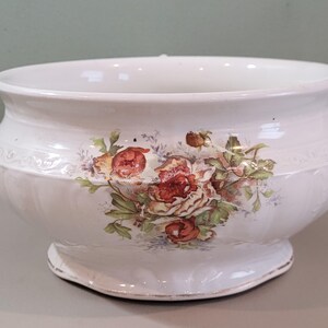 May include: White ceramic planter with a floral design featuring red and pink roses and green leaves. The planter has a wide, shallow bowl shape with a decorative embossed band and a pedestal base.