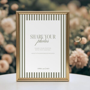 May include: A framed sign with the text "SHARE YOUR photos" in a script font. The sign also includes the text "SHARE YOUR PHOTOS WITH THIS HASHTAG #SMITHTHOMASWEDDING2025" and the names "SOPHIA and JAMES". The frame is gold and the background is white with olive green stripes.