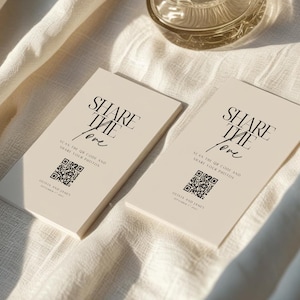 May include: Two cream-colored cards with the text "SHARE THE love" in black. Below the text is a QR code and the words "SCAN THE QR CODE AND SHARE YOUR PHOTOS". The cards are stacked on a cream-colored cloth.
