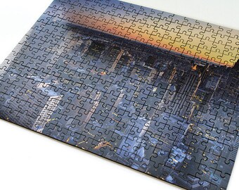May include: A completed jigsaw puzzle showing a cityscape at sunset. The puzzle pieces are arranged to form a panoramic view of a city skyline with tall buildings and a bright orange sky.
