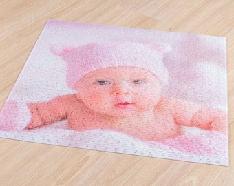 May include: A baby wearing a pink knitted hat and a white onesie is lying on a pink and white blanket. The baby has blue eyes and is looking at the camera.