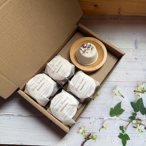 May include: A brown cardboard box containing four wrapped shower steamers and one on a wooden dish. The wrapped steamers have a white label with the text "Shower Steamers Meditation". The unwrapped steamer is white with dried flowers.