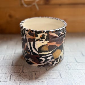 Op de afbeelding: Een cilindervormige keramische plantenpot met een collage-achtig buitenpatroon met zwarte, witte, bruine en beige dierenprints, waaronder luipaardvlekken en zebrastrepen. De binnenzijde van de pot is afgewerkt met een gladde, gebroken witte glazuurlaag.