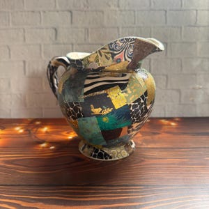 Può includere: Una brocca in ceramica con una superficie in découpage caratterizzata da un collage di vari motivi, tra cui strisce zebrate bianche e nere, accenti in foglia d'oro, piume di pavone color ottanio e una piccola sagoma di uccello dorato.