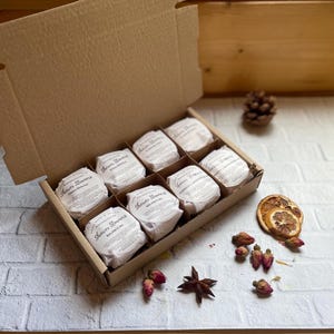 May include: A brown cardboard box containing eight shower steamers, each individually wrapped. The round steamers are wrapped in white paper with labels reading "Shower Steamers". The box is open, revealing the contents. Dried flowers and spices are arranged around the box.