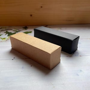 Op de afbeelding: Twee kartonnen dozen, een bruin en een zwart, worden getoond op een wit houten oppervlak. De dozen zijn beide rechthoekig en hebben een eenvoudig ontwerp.