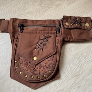 May include: Brown canvas fanny pack with multiple pockets and a belt. The pack features a lace-trimmed pocket, decorative brass studs, and a zippered main compartment. The design includes decorative lacing and brass hardware.