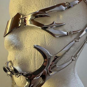 Könnte beinhalten: Eine silberfarbene, avantgardistische Gesichtsmaske mit scharfen, stilisierten Spitzen und Kurven. Die Maske scheint aus poliertem Metall gefertigt zu sein und bedeckt Augen, Stirn und Mund. Sie ist mit filigranen Ketten befestigt.