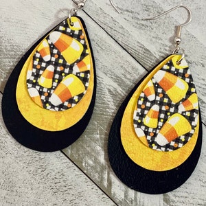 Può includere: Un paio di orecchini pendenti con un design a strati. Lo strato superiore presenta un motivo di mais dolce bianco e nero, lo strato intermedio è giallo e lo strato inferiore è nero. Gli orecchini sono attaccati a ganci argentati.