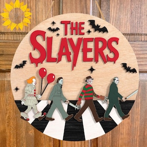 Puede incluir: Un letrero redondo de madera con el texto THE SLAYERS en fuente roja estilo goteo. Debajo, cuatro personajes de películas de terror caminan sobre un camino a rayas blancas y negras. El diseño incluye murciélagos negros, estrellas y dos globos rojos.