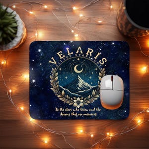Könnte beinhalten: Rechteckiges Mauspad mit Sternennacht-Design und dem Text "VELARIS CITY OF STARLIGHT". Das Design umfasst einen Halbmond, Berge und einen floralen Kranz. Eine weiße und orangefarbene Computermaus liegt darauf.