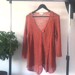Könnte beinhalten: Ein rostfarbener Cardigan mit offenem Vorderteil und einem einzigen Knopfverschluss. Der Cardigan ist aus einem leichten, offen gewebten Stoff gefertigt.