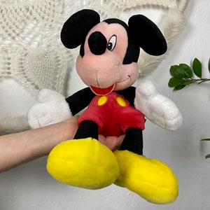May include: A plush Mickey Mouse toy with black ears, a red shirt with yellow buttons, and yellow shoes. The toy is sitting on a person's hand.