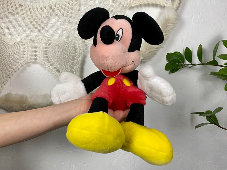 May include: A plush Mickey Mouse toy with black ears, a red shirt with yellow buttons, and yellow shoes. The toy is sitting on a person's hand.