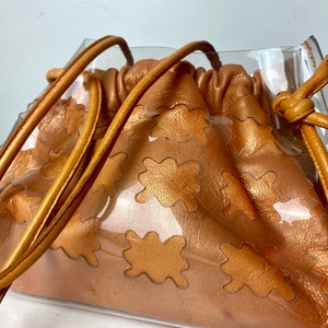 May include: A clear plastic bag with a brown leather drawstring bag inside. The leather bag has a pattern of small, embossed flowers.