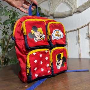 May include: Red backpack featuring Mickey Mouse and star graphics. The backpack has multiple zippered compartments with yellow trim and blue accents. One pocket has the text "Oh Boy!"